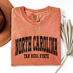 May include: A coral-colored Comfort Colors t-shirt with the words "NORTH CAROLINA" arched across the chest in black block letters, with "TAR HEEL STATE" below. A gold necklace and a brown hat are next to the shirt.