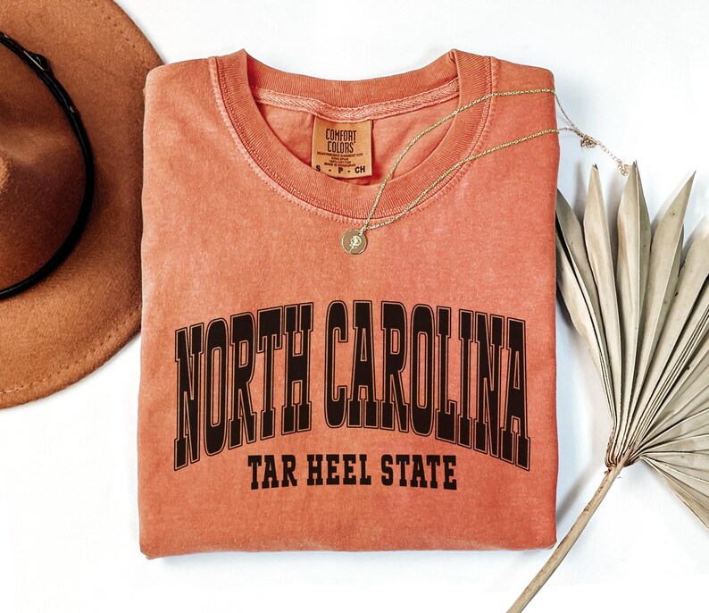 May include: A coral-colored Comfort Colors t-shirt with the words "NORTH CAROLINA" arched across the chest in black block letters, with "TAR HEEL STATE" below. A gold necklace and a brown hat are next to the shirt.
