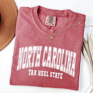 May include: A red Comfort Colors t-shirt with the words "NORTH CAROLINA" and "TAR HEEL STATE" printed in white. A gold necklace with a pendant rests on the shirt. A brown hat and dried leaves are in the background.