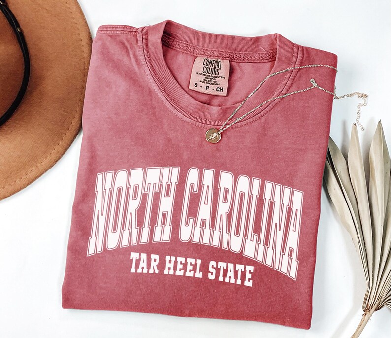 May include: A red Comfort Colors t-shirt with the words "NORTH CAROLINA" and "TAR HEEL STATE" printed in white. A gold necklace with a pendant rests on the shirt. A brown hat and dried leaves are in the background.