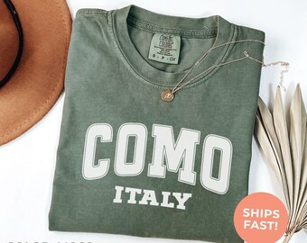 May include: A moss green t-shirt with the word "COMO" in large white block letters above "ITALY". A gold necklace rests on the shirt. The shirt is folded and next to a brown hat and dried leaves.