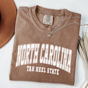 A espresso color shirt, illustrating a varsity-style design with bold “North Carolina” lettering — perfect for cozy, college-inspired style with a touch of the outdoors.