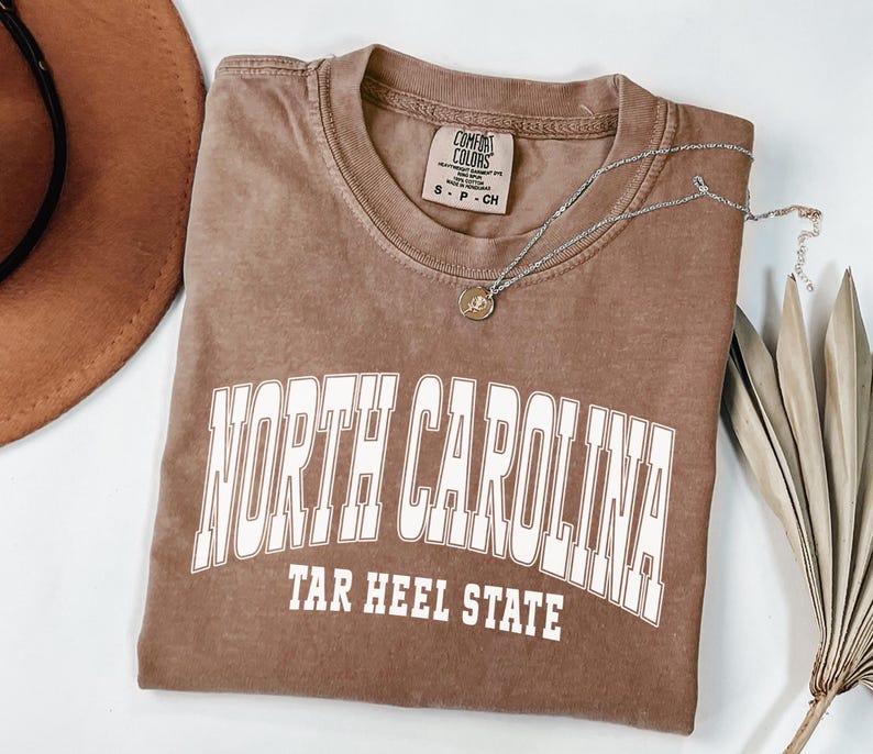 A espresso color shirt, illustrating a varsity-style design with bold “North Carolina” lettering — perfect for cozy, college-inspired style with a touch of the outdoors.