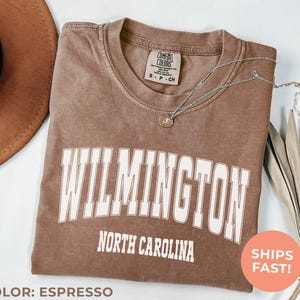 An espresso-colored shirt, illustrating a varsity-style design with bold “Wilmington” city lettering — perfect for cozy, college-inspired style with a touch of the outdoors.