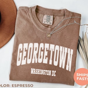 May include: Espresso-colored t-shirt with "GEORGETOWN WASHINGTON DC" printed in white block letters. The shirt has a crew neck and a small tag near the collar. A silver necklace with a pendant rests on the shirt. A brown hat and dried leaves are in the background.