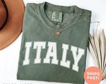 May include: A moss green t-shirt with the word "ITALY" in large white block letters. The shirt has a crew neck and a small tag on the collar. A gold necklace with a pendant rests on the shirt. The text "SHIPS FAST!" is on a round orange sticker.