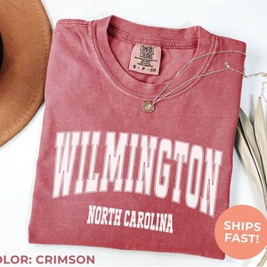 May include: Crimson-colored t-shirt with the word "WILMINGTON" in white block letters, and "NORTH CAROLINA" below. A gold necklace with a pendant rests on the shirt. A brown hat is visible in the upper left corner.