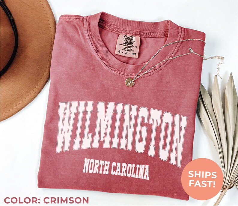 May include: Crimson-colored t-shirt with the word "WILMINGTON" in white block letters, and "NORTH CAROLINA" below. A gold necklace with a pendant rests on the shirt. A brown hat is visible in the upper left corner.