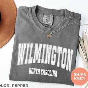 May include: Pepper-colored t-shirt with the word "WILMINGTON" in large white block letters, and "NORTH CAROLINA" below. A gold necklace with a pendant rests on the shirt. The shirt is folded, with a brown hat in the background.