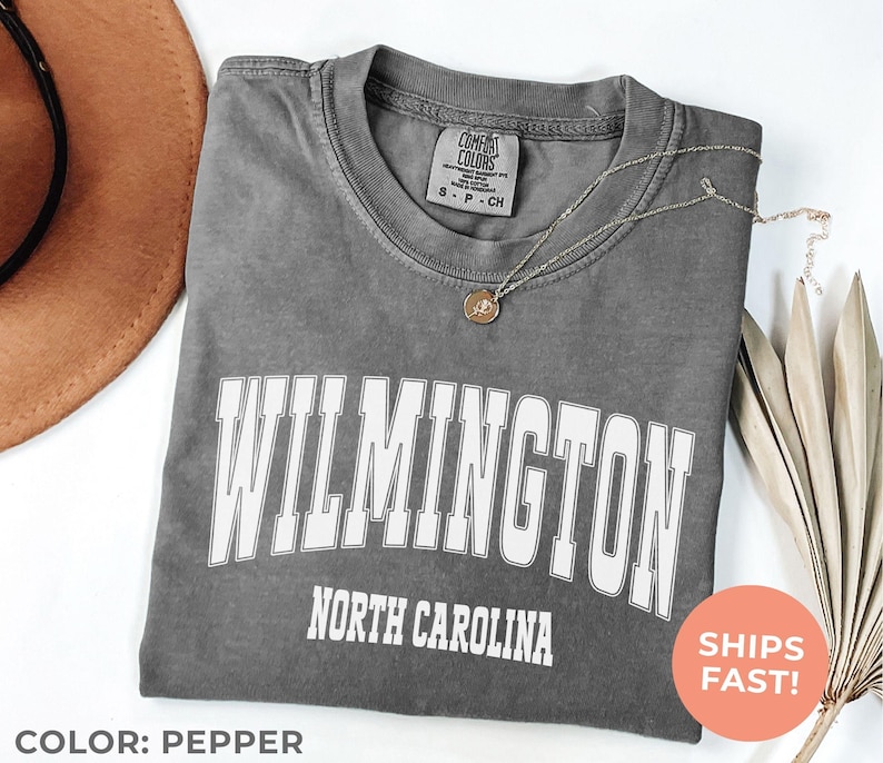 May include: Pepper-colored t-shirt with the word "WILMINGTON" in large white block letters, and "NORTH CAROLINA" below. A gold necklace with a pendant rests on the shirt. The shirt is folded, with a brown hat in the background.