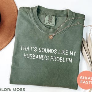 May include: Moss green t-shirt with the text "THAT'S SOUNDS LIKE MY HUSBAND'S PROBLEM" in white. The shirt has a crew neck and short sleeves. A gold necklace with a pendant is on the shirt. The color of the shirt is moss.