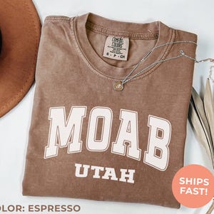May include: Espresso-colored t-shirt with the word "MOAB" in large white block letters, and "UTAH" below. A silver necklace with a small pendant rests on the shirt. The shirt has a Comfort Colors tag. A brown hat is in the upper left corner.