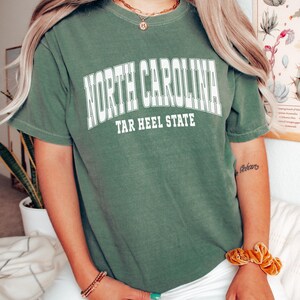 May include: Olive green t-shirt with the text "NORTH CAROLINA TAR HEEL STATE" in white block letters. The shirt has short sleeves and a crew neck. The model is wearing white pants and gold jewelry.