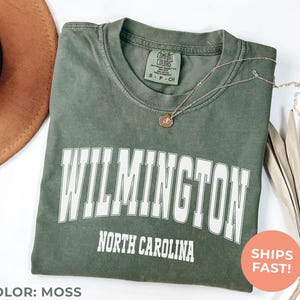 May include: Moss green t-shirt with "WILMINGTON NORTH CAROLINA" printed in white. The shirt has a crew neck and short sleeves. A gold necklace with a pendant is draped across the shirt. A brown hat and dried leaves are in the background.