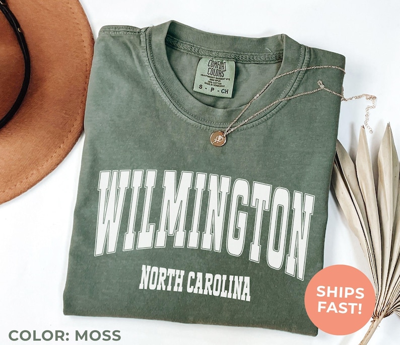 May include: Moss green t-shirt with "WILMINGTON NORTH CAROLINA" printed in white. The shirt has a crew neck and short sleeves. A gold necklace with a pendant is draped across the shirt. A brown hat and dried leaves are in the background.