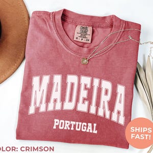 May include: Crimson-colored t-shirt with the word "MADEIRA" in white block letters above "PORTUGAL". A gold necklace with a pendant rests on the shirt. A brown hat and dried leaves are in the background.