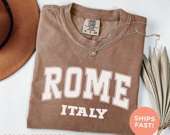 May include: Espresso-colored t-shirt with the word "ROME" in large white block letters, and "ITALY" below. A silver necklace with a pendant rests on the shirt. The shirt has a Comfort Colors tag. A round sticker says "SHIPS FAST!"