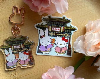 以下が含まれることがあります： 伝統的な衣装を着たウサギとハローキティが描かれた、ローズゴールドのウサギのキーホルダーと2つのアクリルチャーム。チャームとキーホルダーは、ピンクの花が飾られた木製のトレイの上にあります。