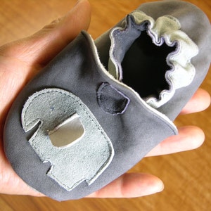 Könnte beinhalten: Ein Paar graue Babyschuhe mit einem hellgrauen Elefanten-Applikationen auf der Vorderseite. Die Schuhe haben einen weißen Besatz am oberen Rand.