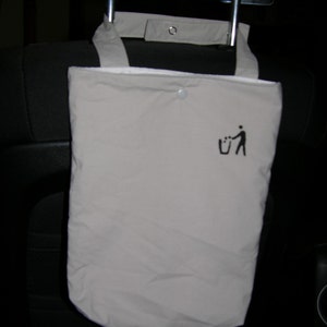 May include: A beige fabric bag with a black trash can and person icon printed on it. The bag is hanging from the headrest of a car seat.