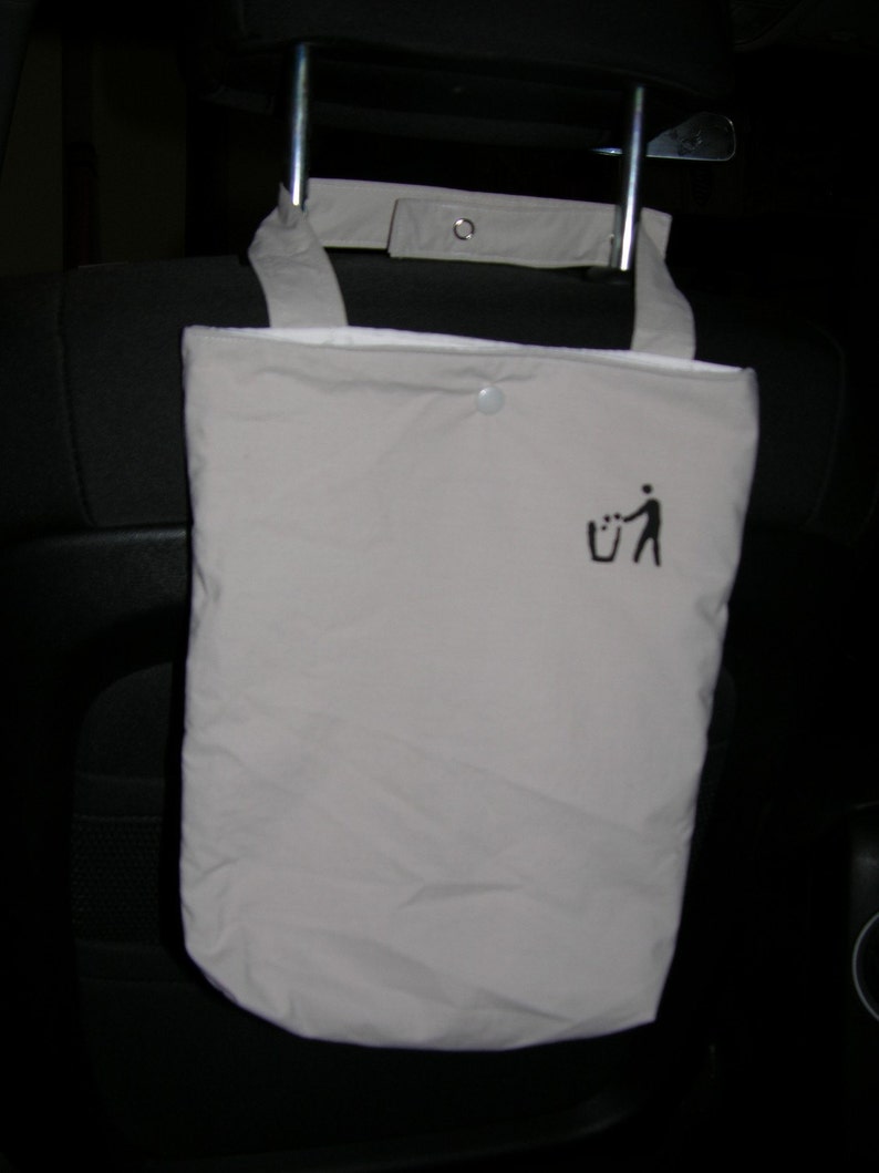 May include: A beige fabric bag with a black trash can and person icon printed on it. The bag is hanging from the headrest of a car seat.