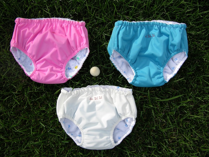 May include: Three pairs of white, pink, and blue swim diapers with a white background and a star pattern. The swim diapers have a white elastic waistband.