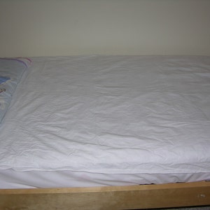May include: A white bed sheet is neatly tucked into a wooden bed frame. A blue pillow with a colorful cartoon character is on the bed.