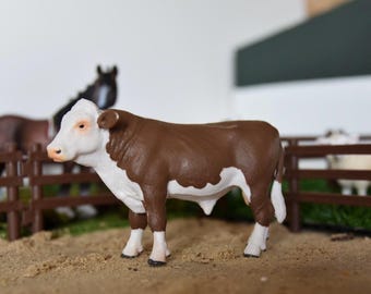 May include: A brown and white toy cow with a white face and a brown body. The cow is standing on a brown wooden fence with a green grassy area behind it.