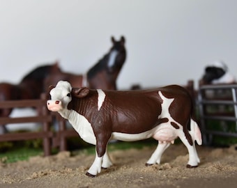 May include: A brown and white plastic toy cow with black hooves and a pink udder. The cow is standing on a brown surface with a wooden fence in the background.