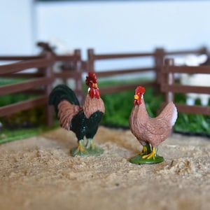 Op de afbeelding: Twee speelgoedboerderijdieren, een haan en een hen, staan op een bruin oppervlak. De haan is zwart en groen met een rode kam en rode keellapjes. De hen is bruin met een rode kam en rode keellapjes.