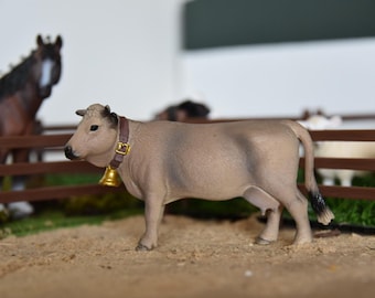 May include: A light brown toy cow with a brown leather collar and a gold bell. The cow is standing on a brown wooden fence with a green grassy area in the background.