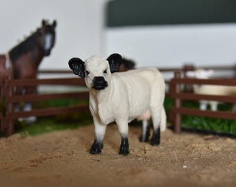 May include: A white and black toy calf stands in a farm setting with a brown fence and a brown horse in the background. The calf has black ears, a black nose, and black hooves. The scene is set on a sandy surface.