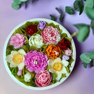 May include: A round, white dish filled with colorful, handcrafted soap flowers. The arrangement includes pink, white, orange, and red blooms, with green leaves. The soap flowers are arranged in a circular pattern, creating a decorative display.