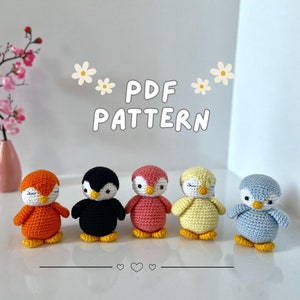 May include: Five crocheted penguin amigurumi toys in different colors: orange, black, pink, yellow, and blue. The toys are arranged in a row on a white surface. The text "PDF PATTERN" is displayed above the toys.
