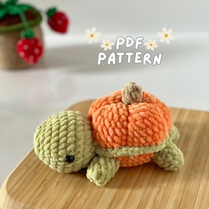 Può includere: Un giocattolo amigurumi a forma di tartaruga zucca lavorato a uncinetto con un guscio verde e un guscio di zucca arancione. Il giocattolo è appoggiato su una superficie di legno. Il testo "PDF PATTERN" è scritto in lettere bianche su uno sfondo bianco con fiori bianchi.