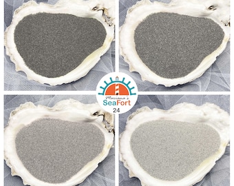 Peut inclure: Quatre coquilles d'huître blanches remplies chacune d'une nuance différente de sable fin, allant du gris anthracite foncé au gris argenté clair. Un logo circulaire au centre affiche une icône de phare et le texte Mariana's SeaFort 24.