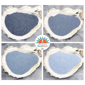 Peut inclure: Quatre coquilles d'huître contenant chacune une nuance différente de sable bleu, allant du bleu marine foncé au bleu ciel clair. Un logo circulaire au centre présente un phare avec le texte Mariana's SeaFort 33 en bleu sarcelle et orange.