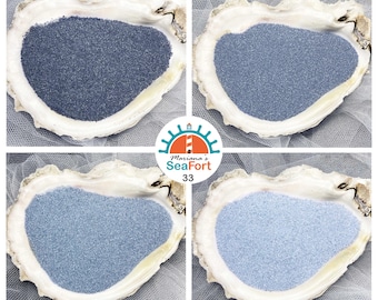 May include: Four oyster shells each containing a different shade of blue sand, ranging from dark navy to light sky blue. A circular logo in the centre features a lighthouse graphic with the text Mariana's SeaFort 33 in teal and orange.