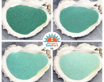 Pode incluir: Quatro conchas de ostra brancas, cada uma preenchida com um tom diferente de areia verde, variando do verde floresta escuro ao verde menta claro. Um logótipo circular no centro apresenta um farol com o texto Mariana's SeaFort 19.