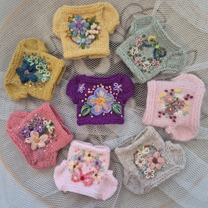 May include: A collection of seven miniature knitted jumpers in various colours including yellow, green, purple, pink, and beige. Each jumper features intricate floral embroidery on the front, showcasing detailed petals, leaves, and stems in vibrant thread.