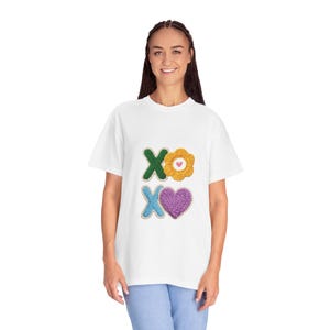 May include: White t-shirt with colorful embroidered designs. The shirt features a green "X" and a yellow flower, and a blue "X" and a purple heart. The design is centered on the front of the shirt.