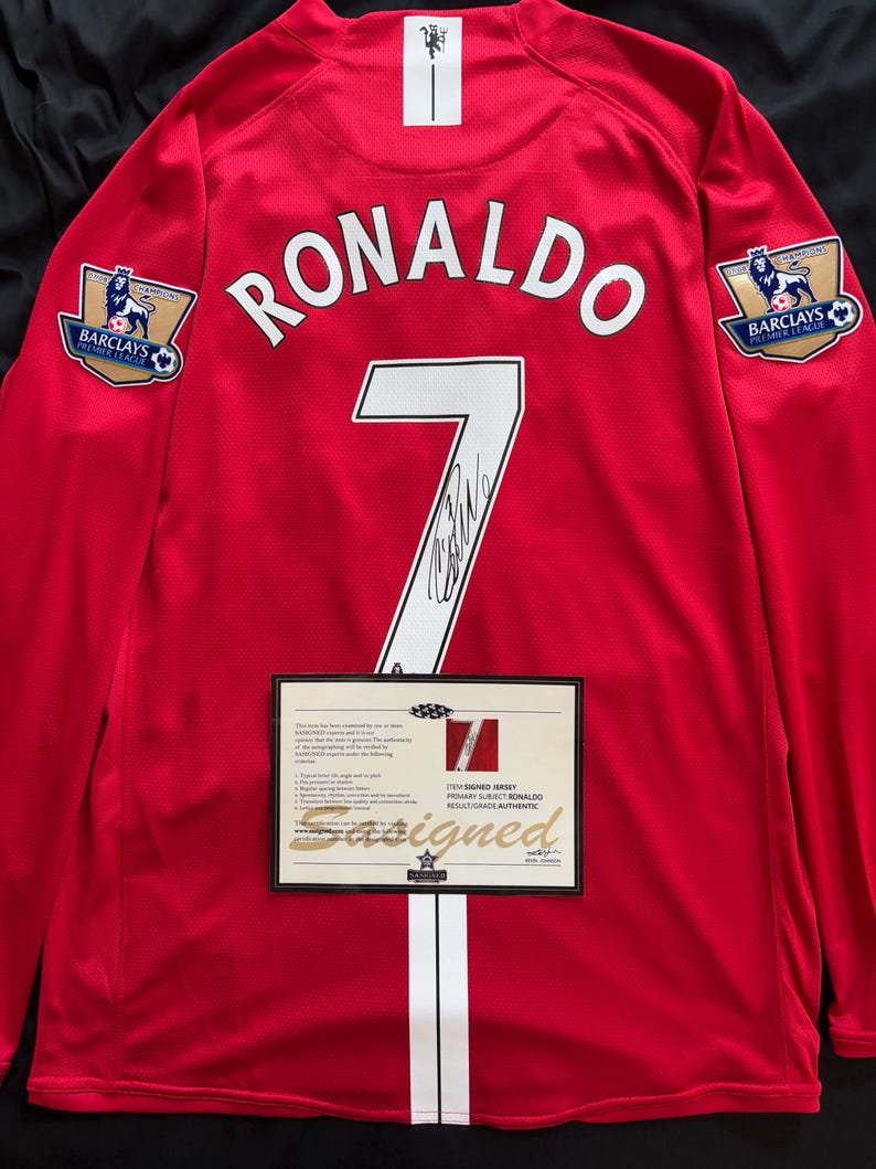 Puede incluir: Camiseta de f&uacute;tbol roja de manga larga con el nombre "RONALDO" y el n&uacute;mero "7" en blanco. Incluye los logotipos de la Premier League Barclays y un certificado de autenticidad. Una franja blanca recorre la parte delantera.