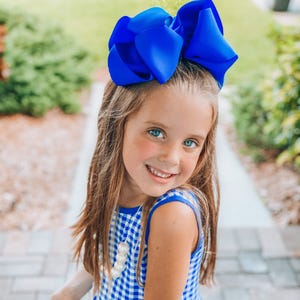 May include: A young girl with long blonde hair wearing a blue and white gingham dress with a blue bow in her hair. She is wearing a white pearl necklace.
