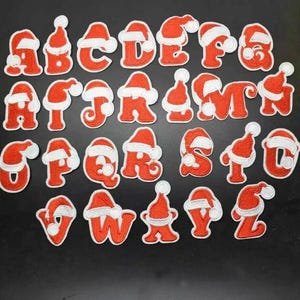 May include: A complete alphabet of red embroidered letters, each decorated with a white-trimmed Santa hat. The letters are in a sans-serif font and are set against a dark background. Suitable for festive crafts and customisation.