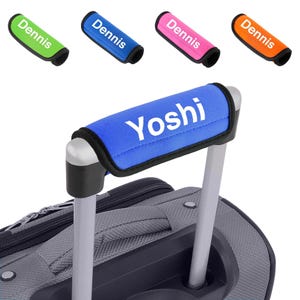May include: A selection of luggage handle wraps in green, blue, pink, and orange, each with the name "Dennis". A blue wrap with "Yoshi" is on a grey suitcase handle. These wraps are designed to personalise and protect luggage handles.