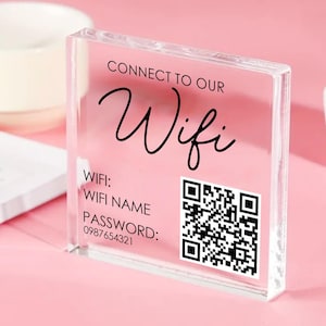 May include: A clear acrylic block with a pink interior, displaying the words "CONNECT TO OUR Wifi" in black script. Below, it lists "WIFI: WIFI NAME" and "PASSWORD: 0987654321" with a QR code. The block is on a pink surface.