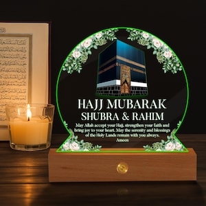 May include: An illuminated sign with the text "HAJJ MUBARAK SHUBRA & RAHIM" and an image of the Kaaba. The sign is set on a wooden base with a warm glow. A lit candle and a book are in the background. The sign is a religious item.