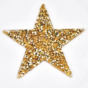 May include: A gold star-shaped embellishment, covered in small, sparkling golden rhinestones. The star has five points and is set against a plain white background, creating a striking contrast.