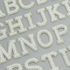 May include: Close-up of white alphabet letters, each encrusted with small, round, pearl-like beads. The letters are arranged on a light grey surface. The letters are capital letters, and the image is well-lit.