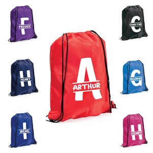 May include: A selection of drawstring bags in various colours, including red, purple, blue, black, and pink. Each bag features a large white initial with a name printed below. The bags are designed for carrying items and have black drawstrings.